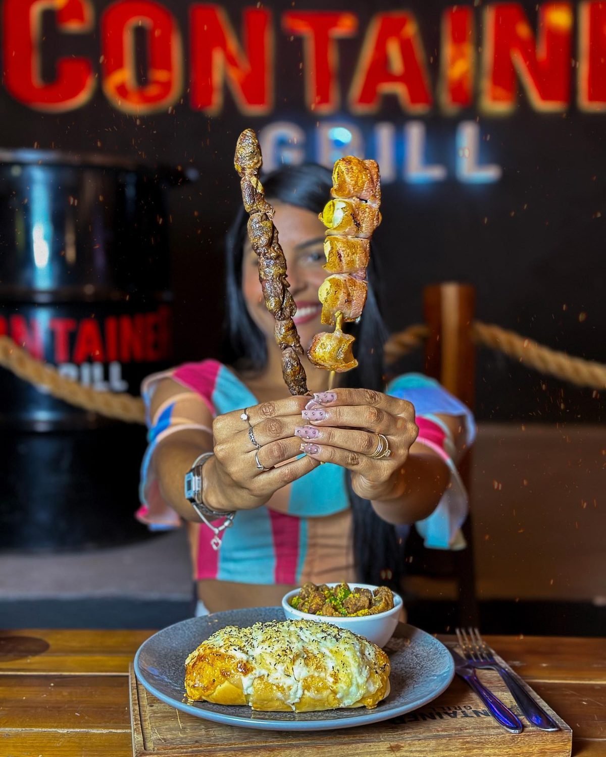 Cliente segurando espetos de carne e queijo em frente ao letreiro iluminado do Container Grill