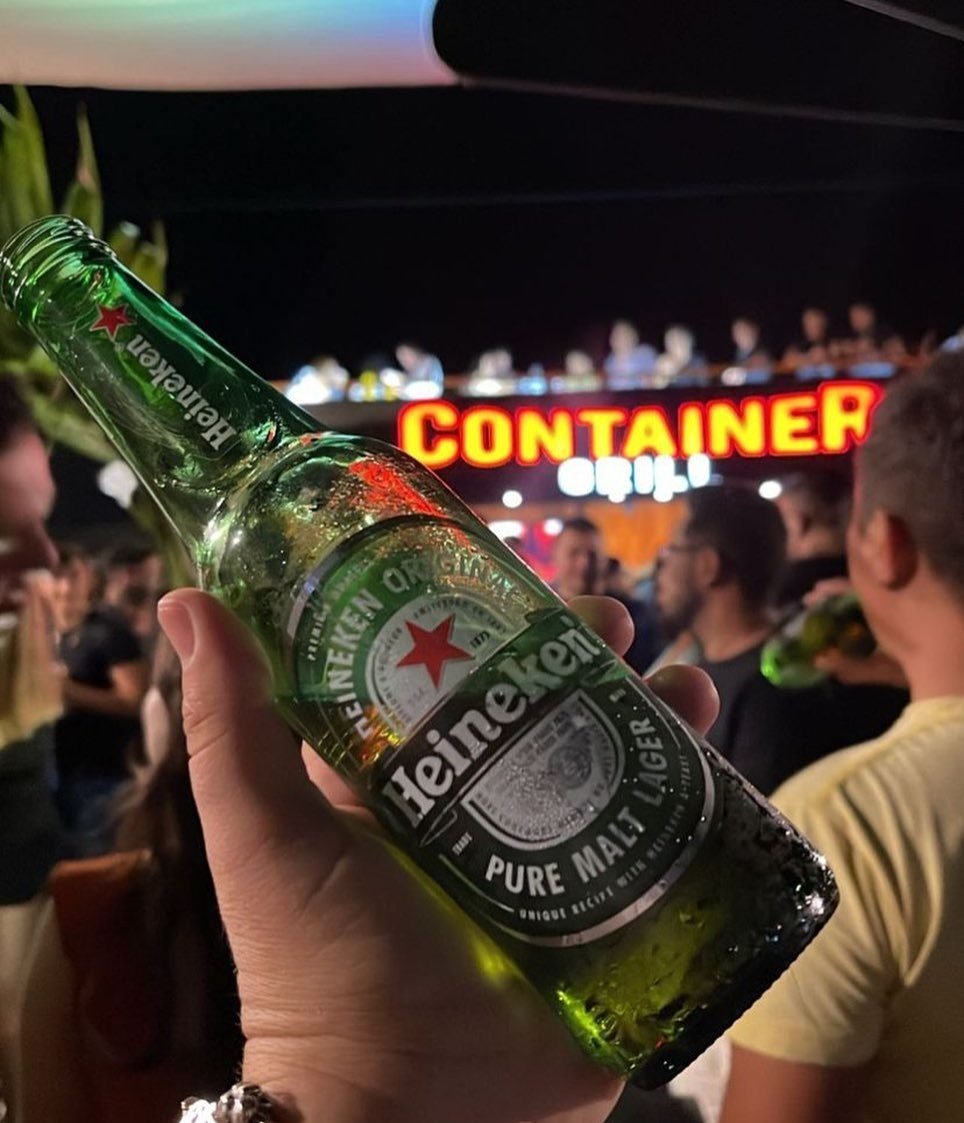 Cerveja erguida na frente do letreiro aceso do Container Grill, com o público ao fundo